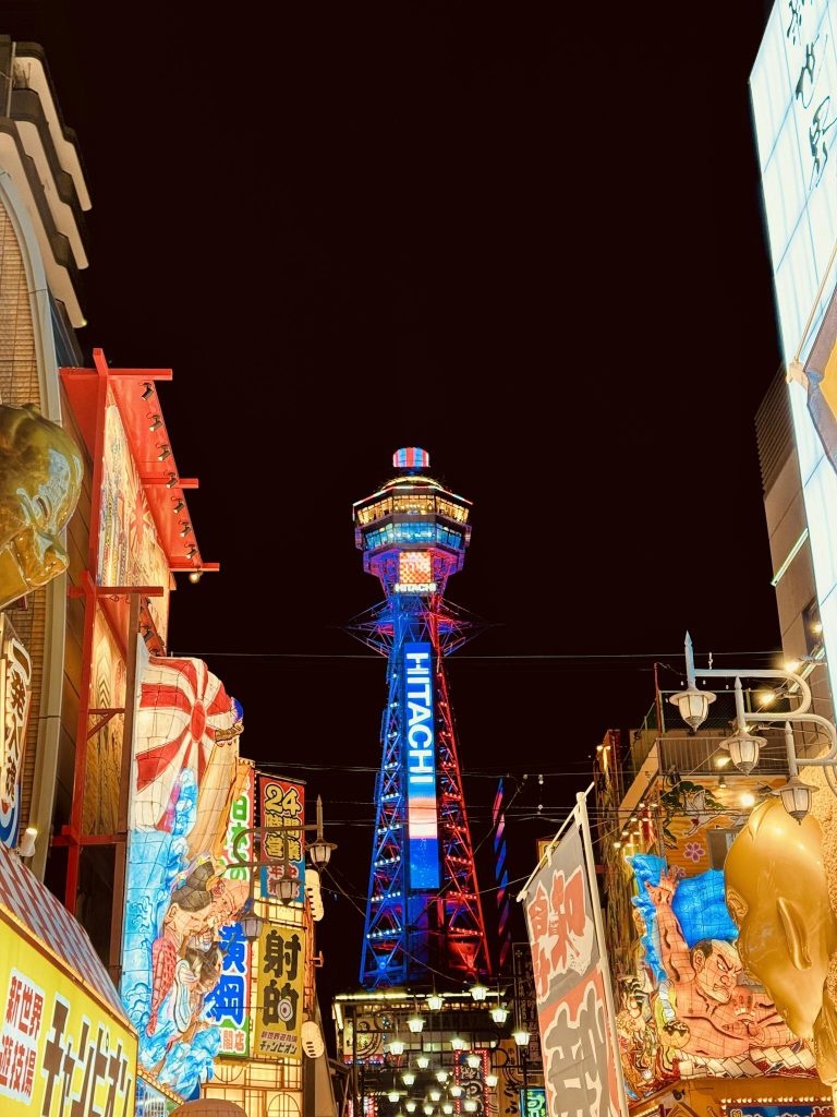 Colorful night view of Tsutenkaku Tower in Osaka, Japan surrounded by neon signs and bustling street life.